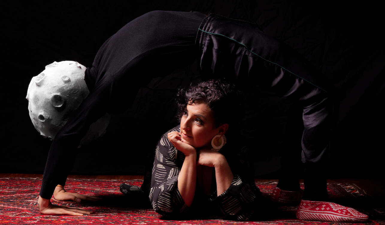 Ayelet Rose Gottlieb laying on the floor in a black and white dress, with someone dressed in black and with a moon head on stretched over her