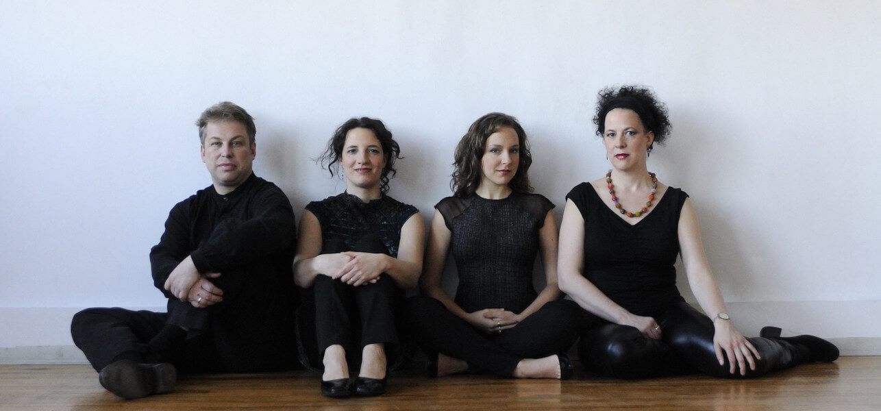 Bozzinni Quartet all in black clothing, sitting on the floor in front of a white wall.