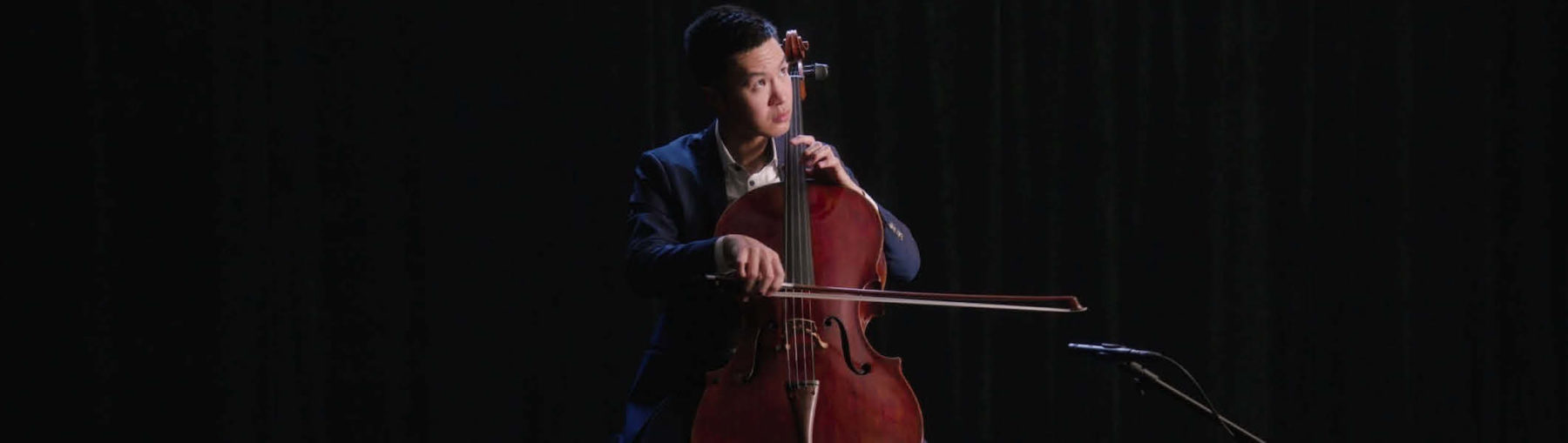 Jonathan Lo in a blue suit sitting behind his cello, looking up.