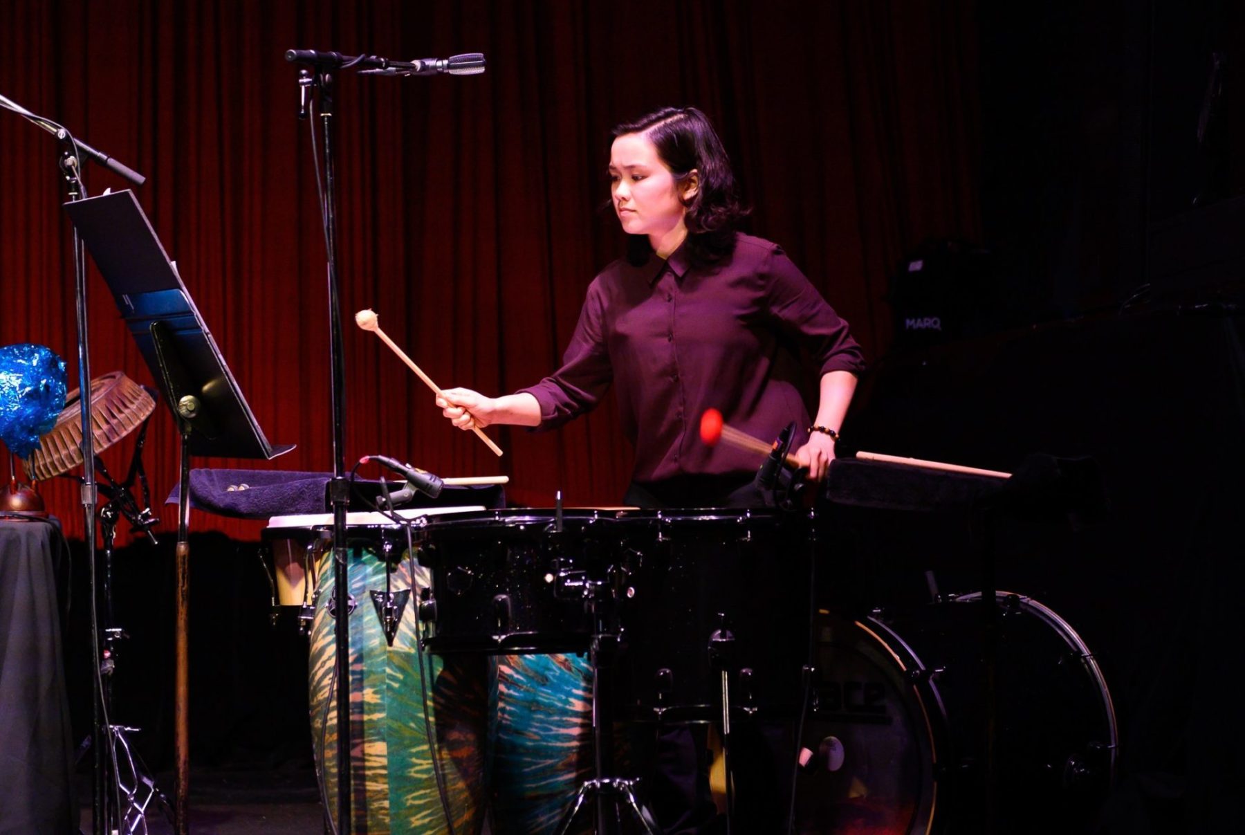 Julia Chien in a purple shirt holding two mallets and playing the drums