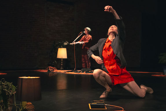 Caroline Shaw in the background singing with Vanessa Goodman in an orange jumper dancing in the foreground