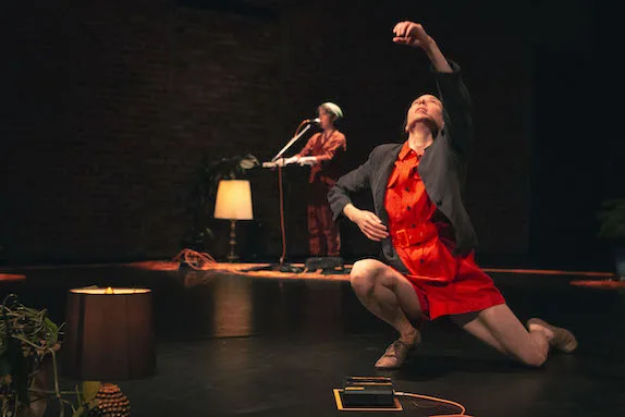 Caroline Shaw in the background singing with Vanessa Goodman in an orange jumper dancing in the foreground