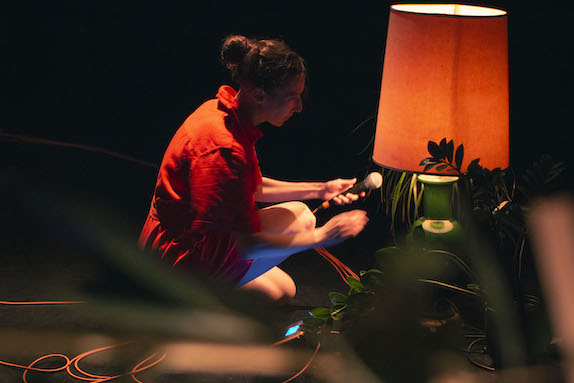 Vanessa Goodman in an orange jumper holding a microphone by a lamp and plants