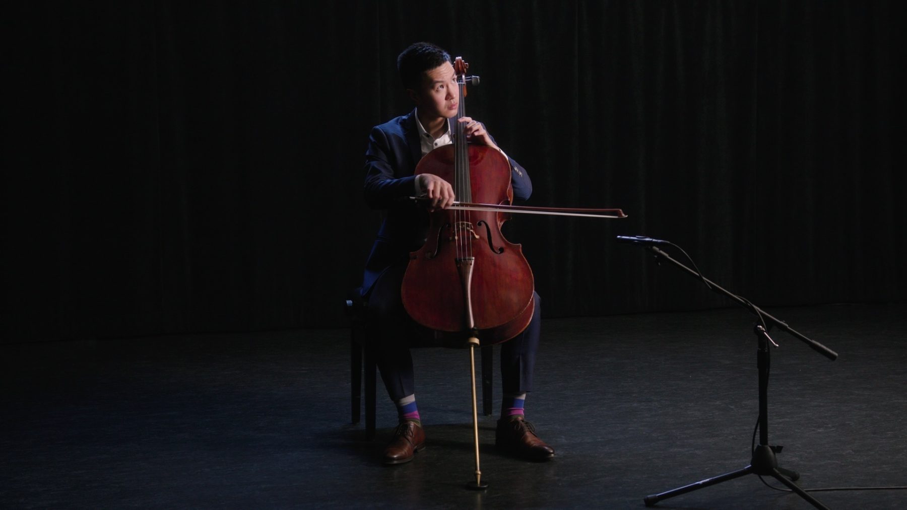 Jonathan Lo in a blue suit sitting behind his cello, looking up.