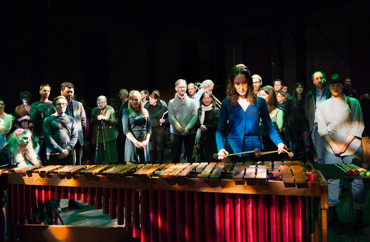 Katie Rife in a blue shirt looking down and playing the vibraphone with four mallets, with audience standing behind her
