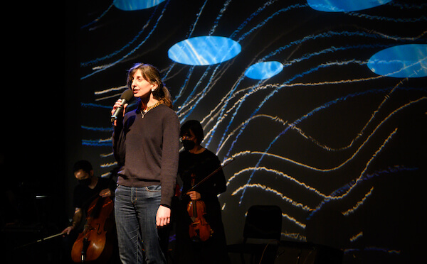 Barbara Alder standing with a microphone in a black shirt and jeans, in front of a screen with blue ovals and lines