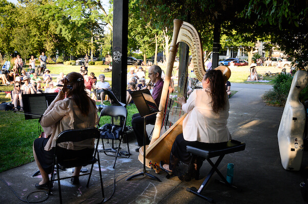Min Jee Yoon with her back to the camera, Lani Krantz with her harp and Paolo Bortolussi performing in the park