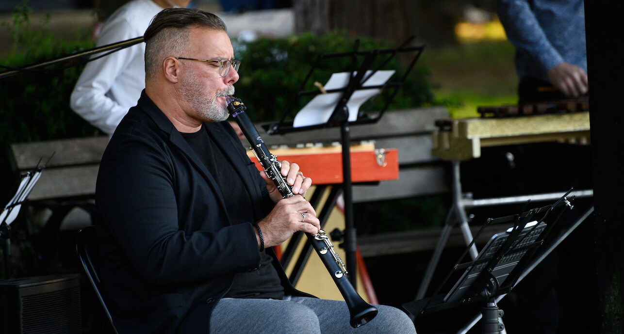 François Houle in glasses, black jacket and grey pants playing the clarinet