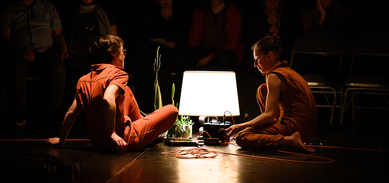Caroline Shaw and Vanessa Goodman sitting by a plant and a turned on lap in a dark theatre with audience slightly seen in the background