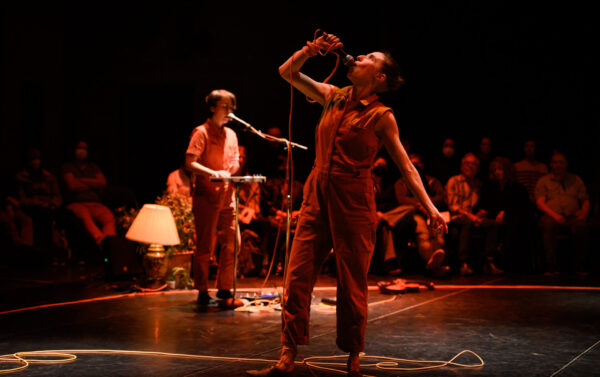 Caroline Shaw in the background singing and playing music with Vanessa Goodman in the foreground speaking into a microphone she holds high up