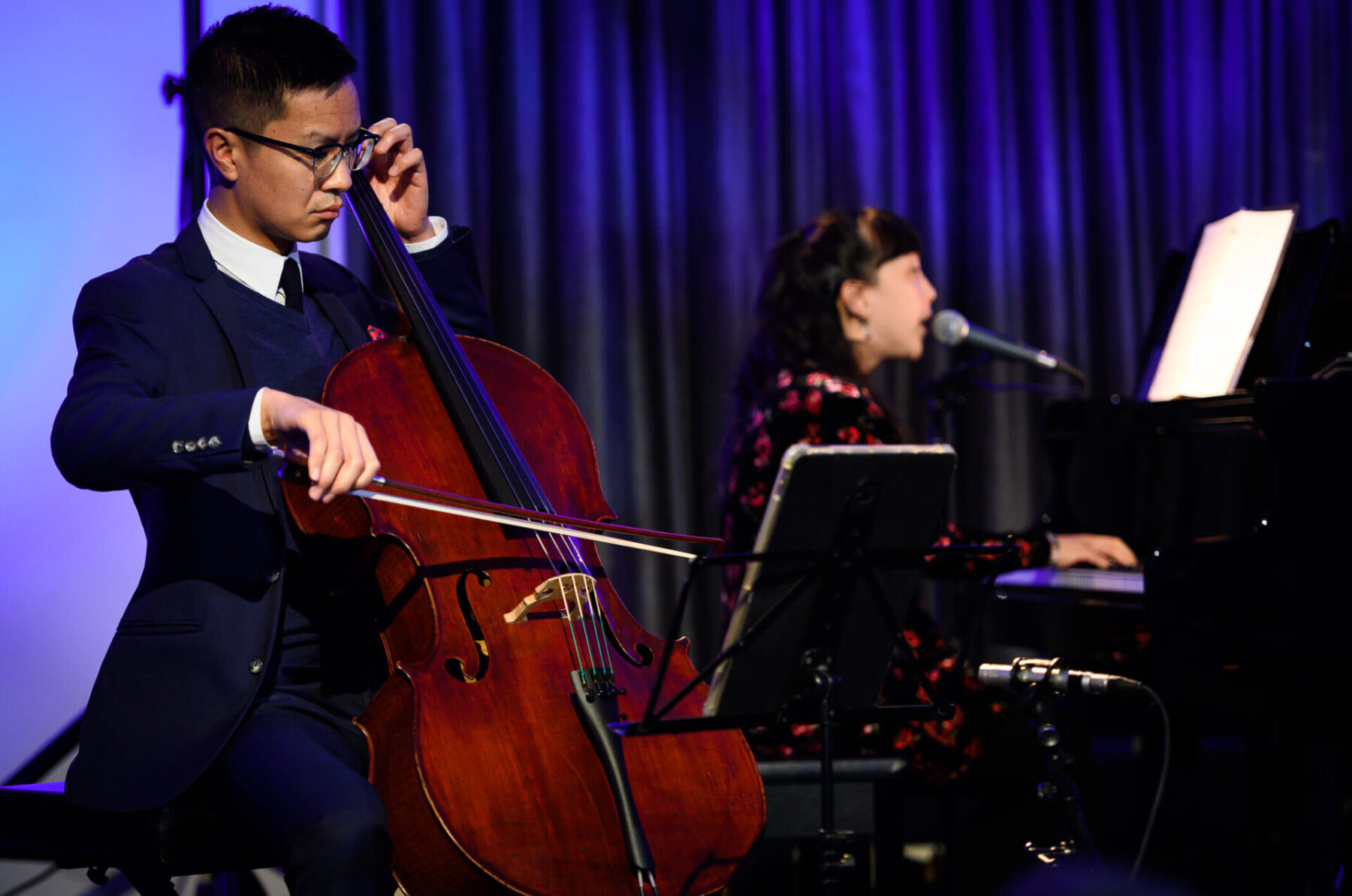 Jonathan Lo in a blue suit on the cello with Robyn Jacob at the piano in the background