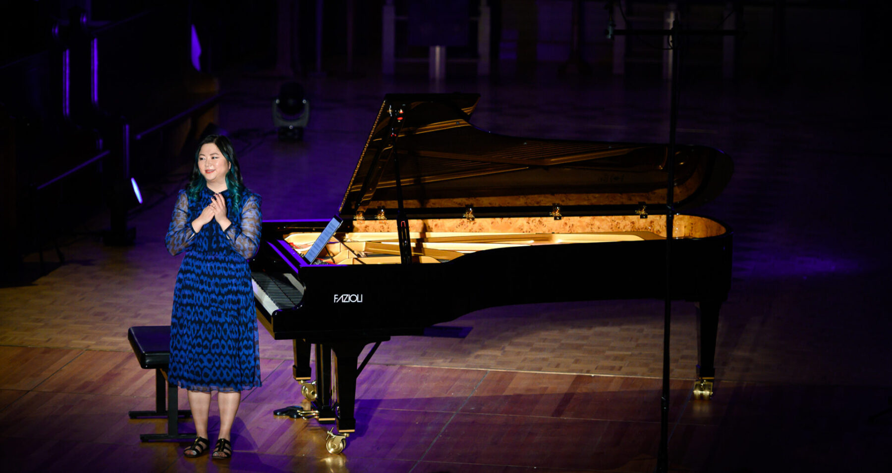 Vicky Chow in a blue and black dress in front of a Fazioli piano, talking to the audience