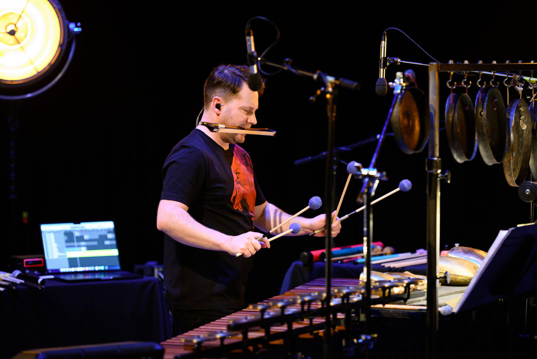 Joby Burgess wearing a black and red shirt with a string bow in his mouth, playing percussion