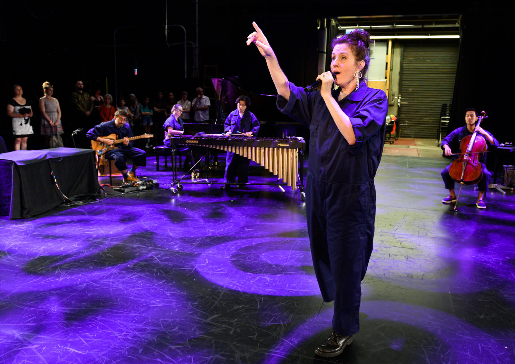 Julia standing and pointing upwards while on the Playhouse stage, lit in purple light.