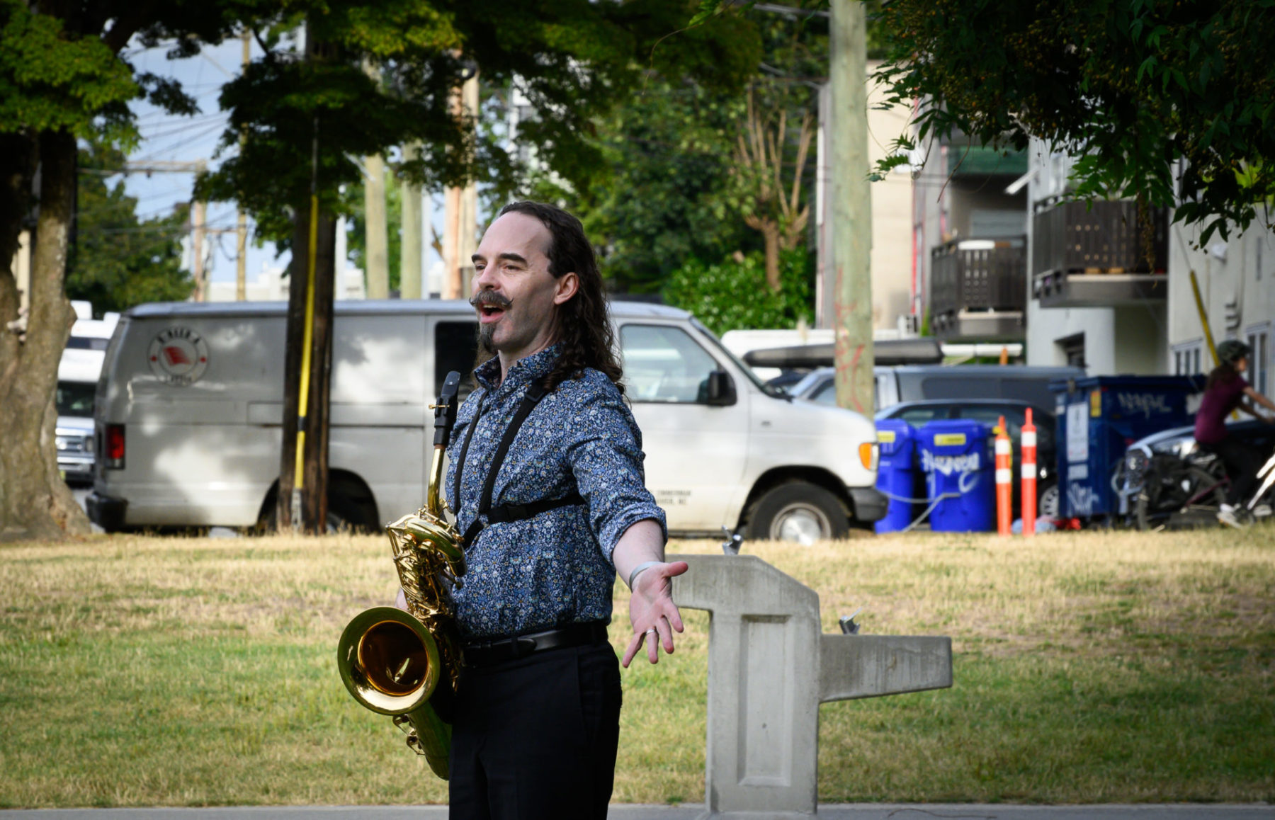 Colin MacDonald in a blue shirt with his saxophone speaking at the park performance
