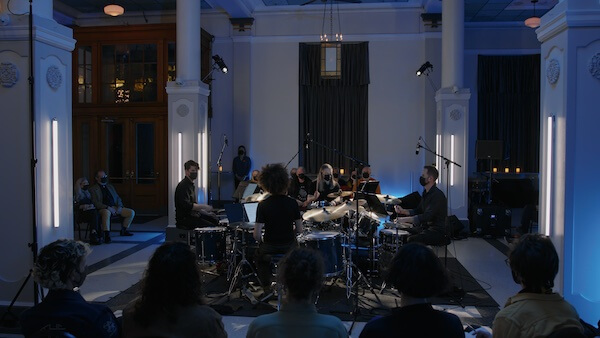 Architek Percussion sitting in a square with black clothes and masks on, with the audience sitting around them