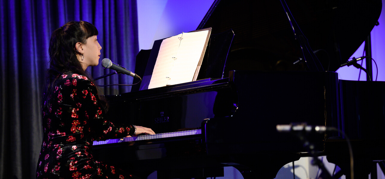 Robyn Jacob in a black dress with red flowers sitting at the piano, back lit with purple light.