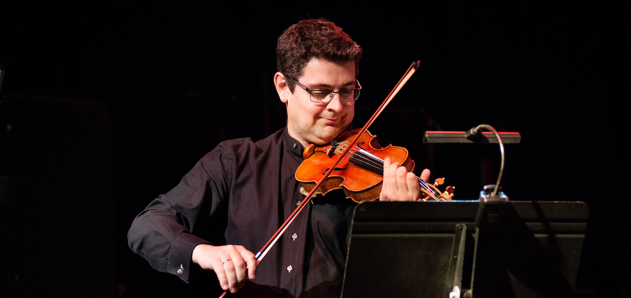 Nicholas Wright standing at a music stand, playing violin, wearing a black button up.