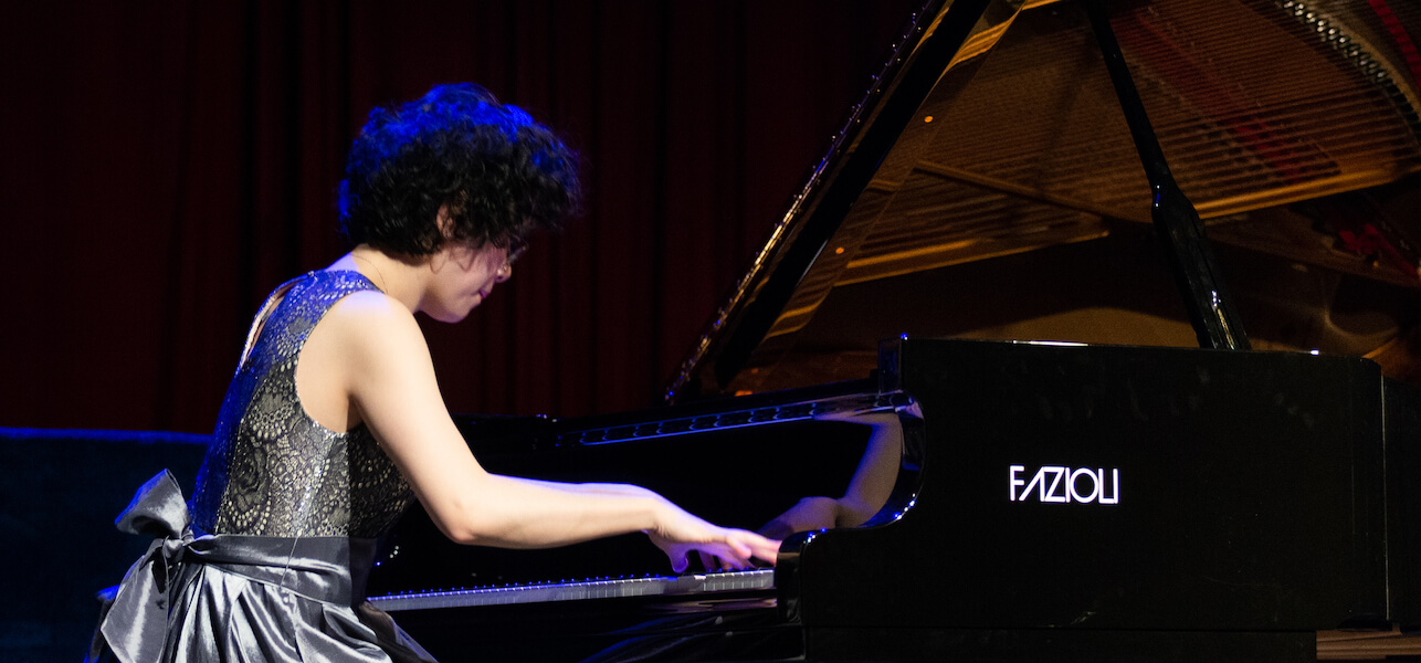 Nicole Linaksita, wearing a grey dress with lacy top, playing a piano with a purple light on her.