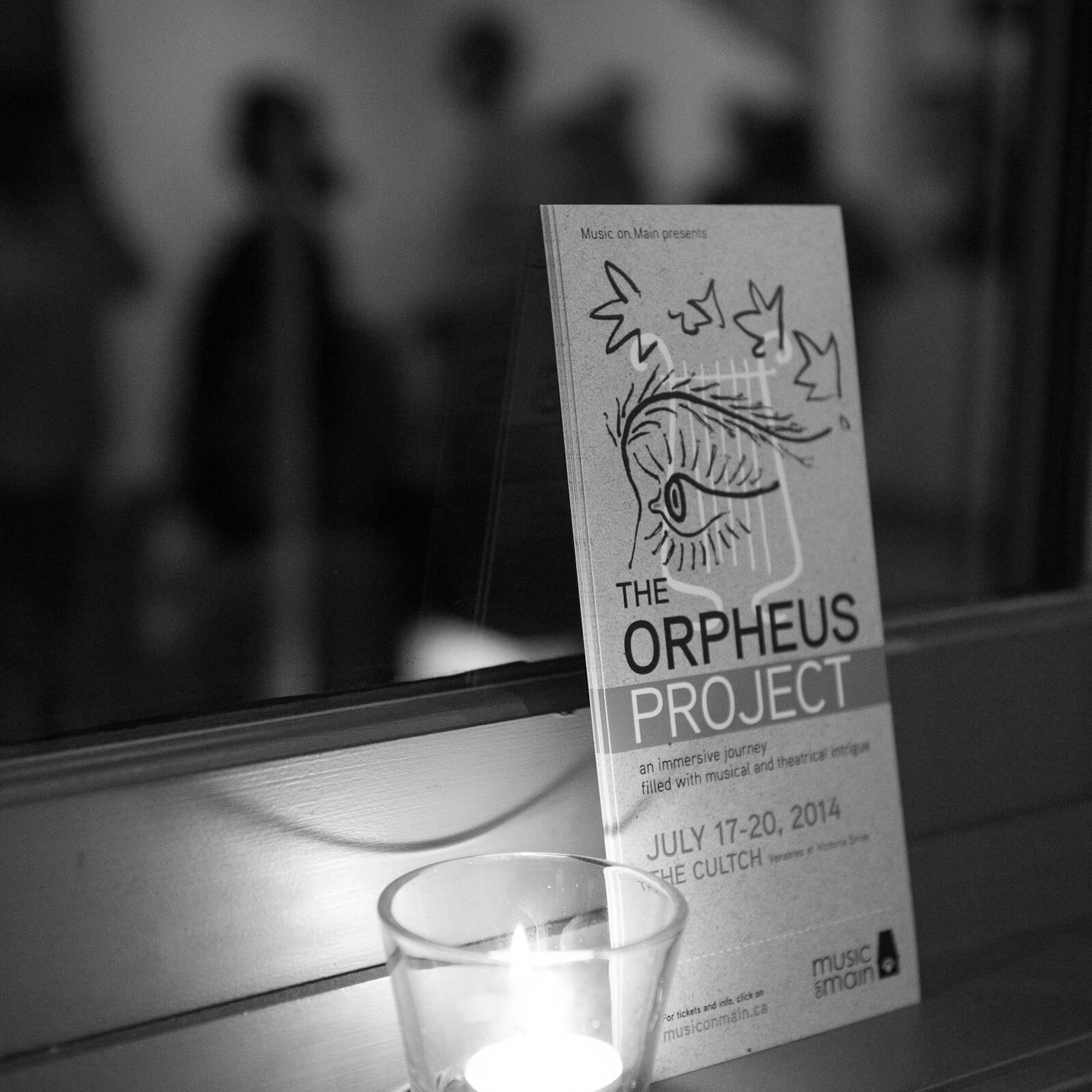 The Orpheus Project postcard leaning against a window with a candle beside it.