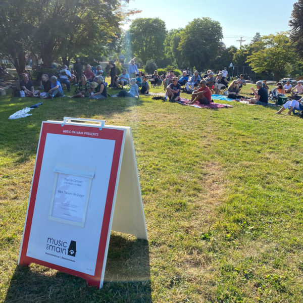 sandwich board that says "pop up concert with Music on Main" in Mount Pleasant Park with audience members sitting on the grass