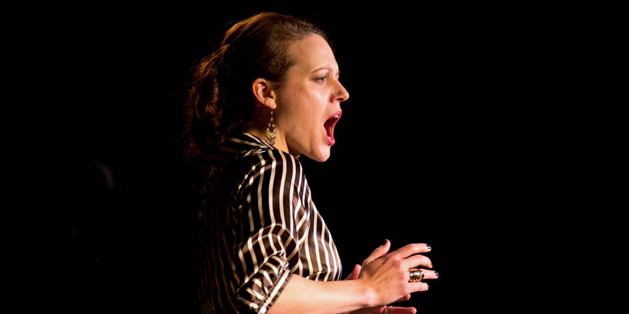 A profile photo of Robyn Driedger-Klassen, wearing a black and white striped shirt and dangling earrings, singing.