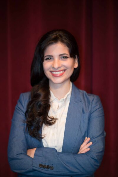 Smiling brightly, Sara is in a white blouse and blue jacket, in front of a red curtain.