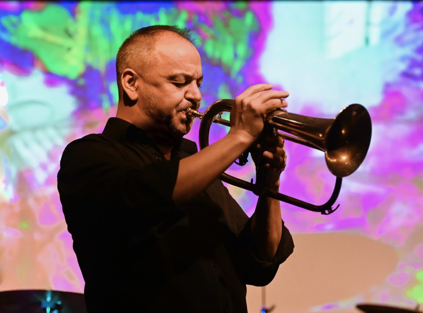 John Korsrud in a black shirt playing the trumpet in front of a multi-coloured screen