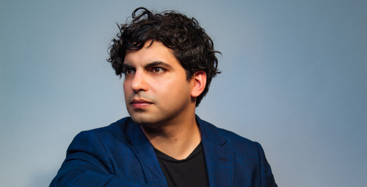 Zubin Kanga in a black shirt and dark blue jacket, looking away from the camera, against a blue background.