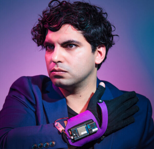 Zubin Kanga wearing a dark blue jacket and electronic glove, in front of a purple ombre background.