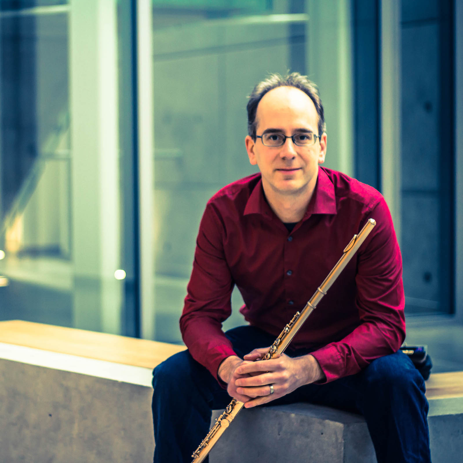 Paolo Bortolussi, in a red shirt and black pants, holding his flute while sitting on a bench in front of a window